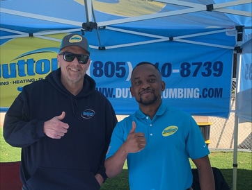 Two smiling men in company shirts giving thumbs up under a branded plumbing tent at an outdoor event