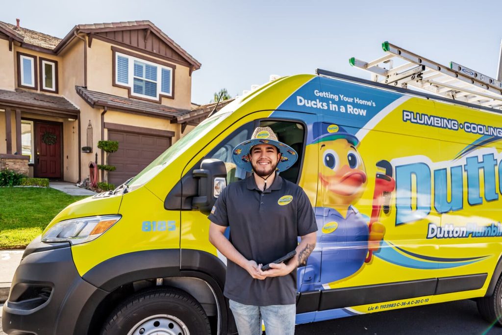 Smiling plumber in hat holding a tablet beside a yellow Dutton Plumbing service van with duck mascot.