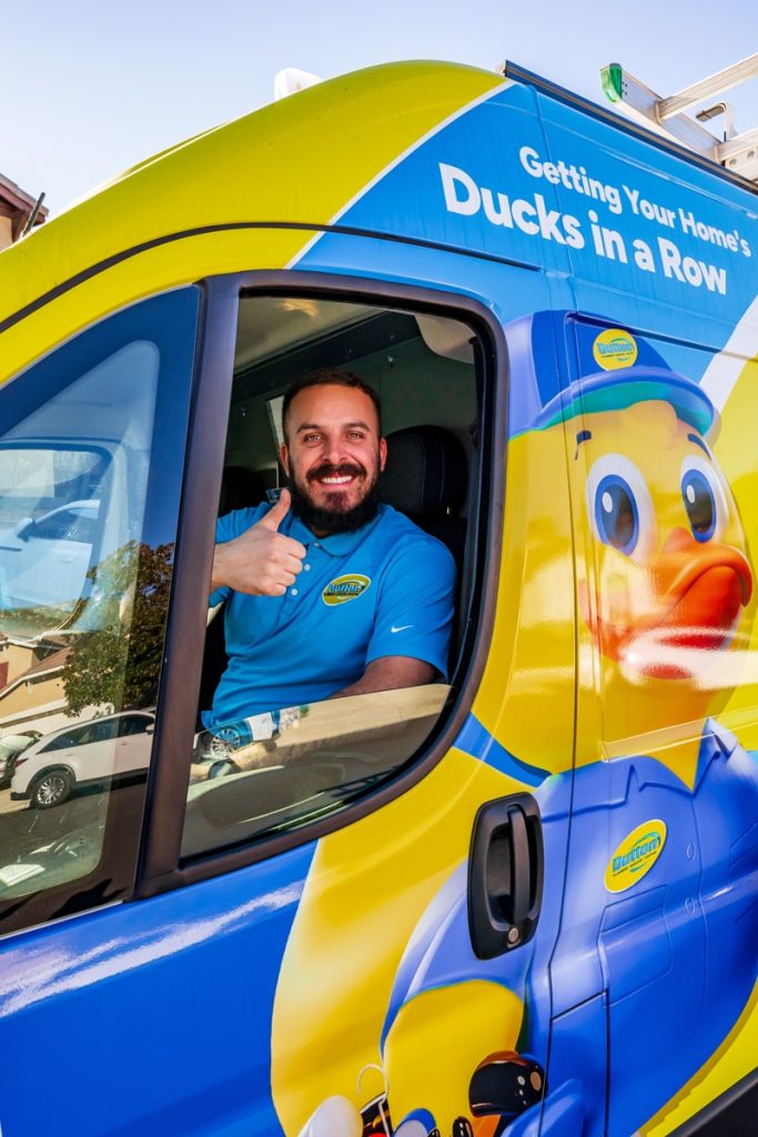 Smiling service technician giving thumbs-up from driver's seat of bright yellow-blue duck-branded service van