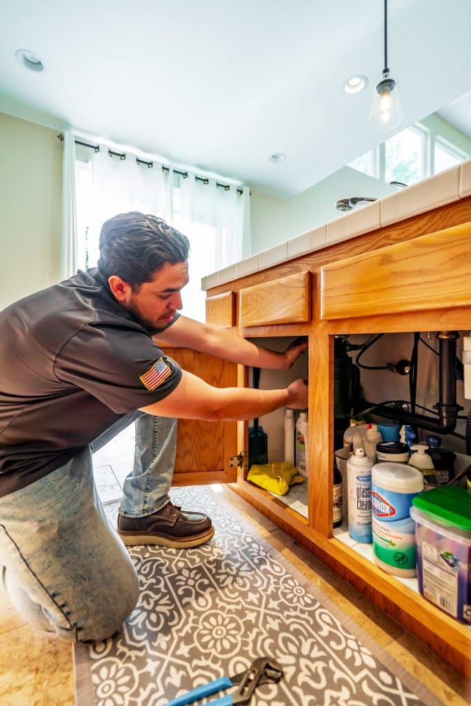 Plumber repairing kitchen sink pipes under a wooden cabinet, kneeling on patterned rug