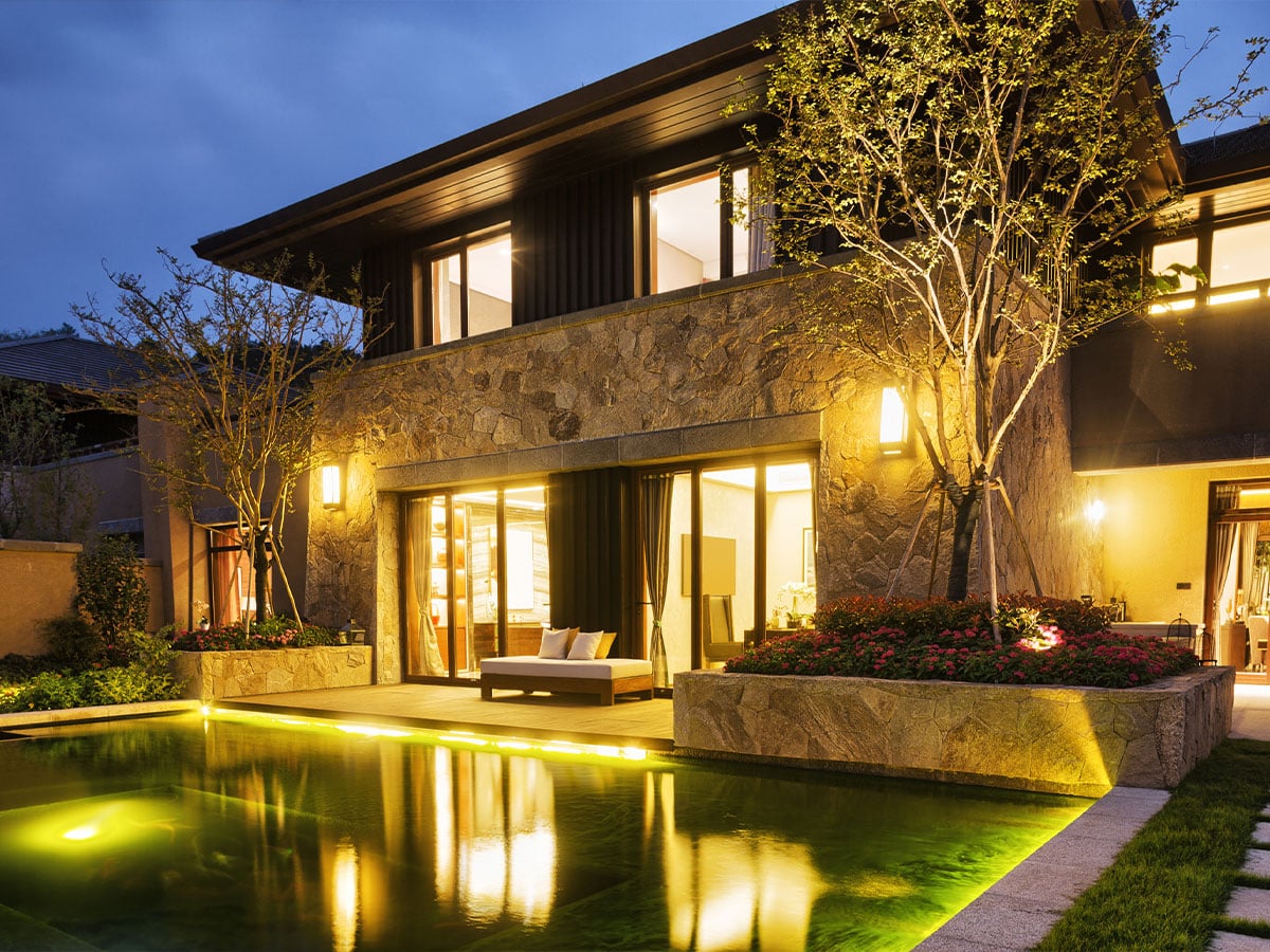 Luxury modern stone house at dusk with warm lights, patio seating and an illuminated swimming pool reflecting landscaping.