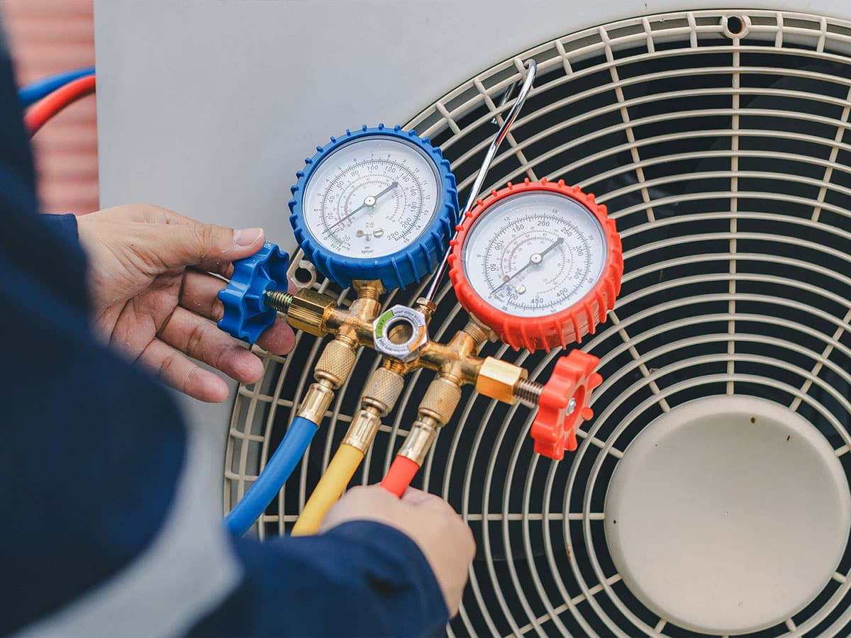 A technician attaches blue and red manifold pressure gauges to an outdoor air conditioner unit for HVAC servicing.