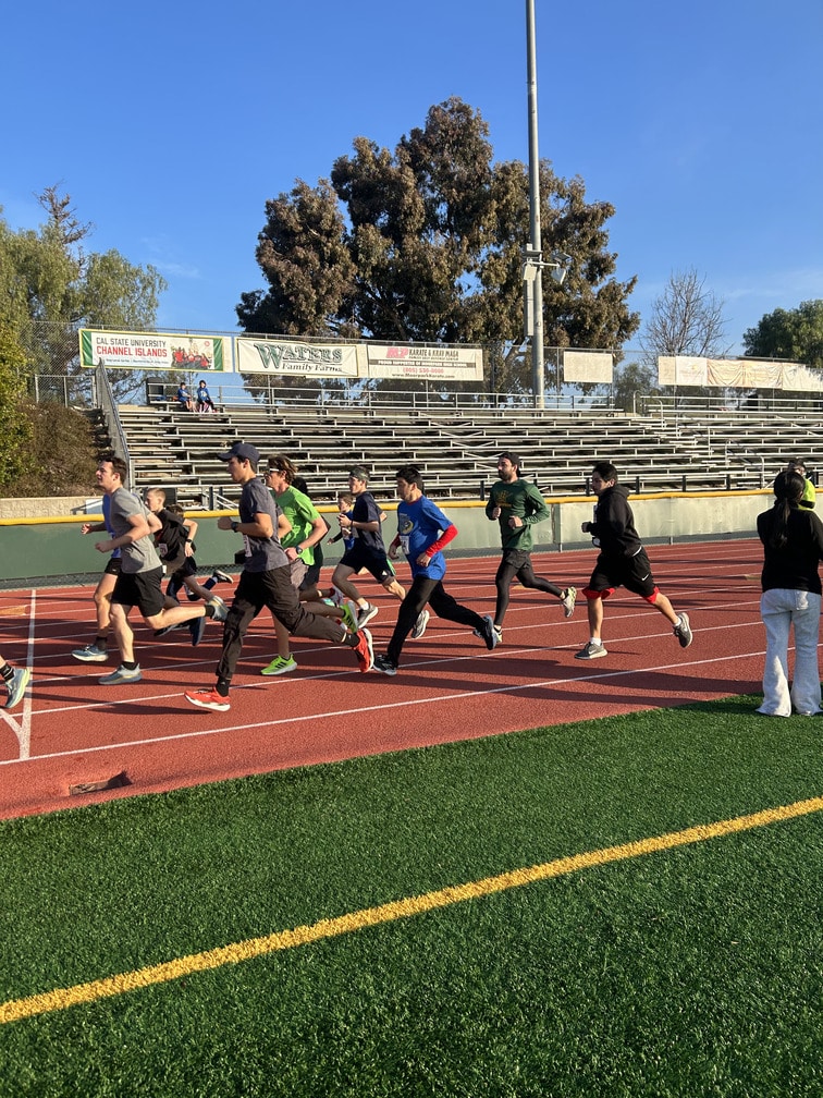 Group of runners sprinting on a red track at a school stadium with empty bleachers and clear blue sky