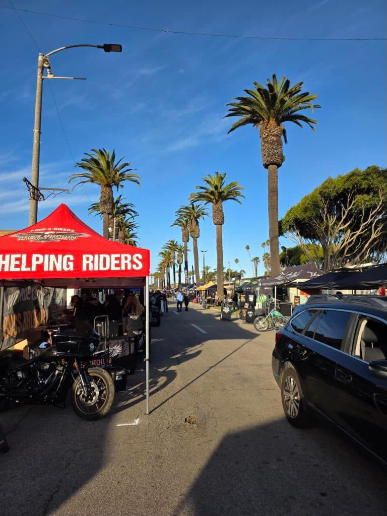 Outdoor motorcycle market on palm-lined street with 'Helping Riders' red tent, bikes, vendors and clear blue sky