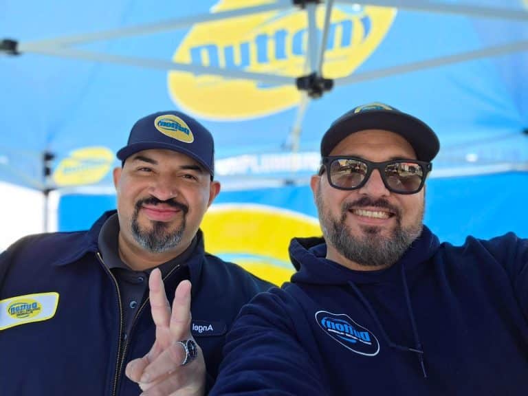 Two smiling men in branded blue caps and jackets taking selfie under blue-and-yellow promotional canopy