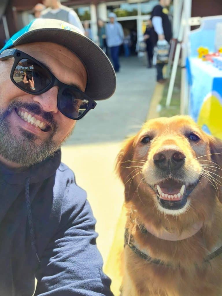 A man in sunglasses and a cap takes a selfie with a smiling golden retriever at an outdoor event