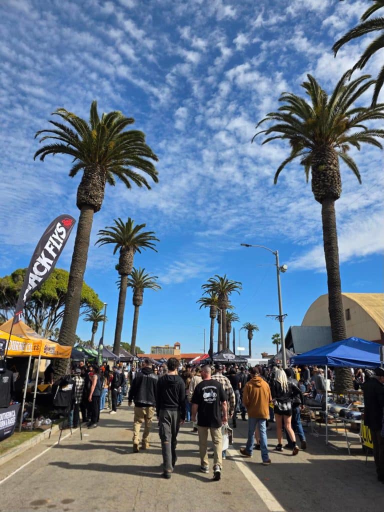 People walking through a street fair lined with palm trees and vendor tents under a bright blue sky.