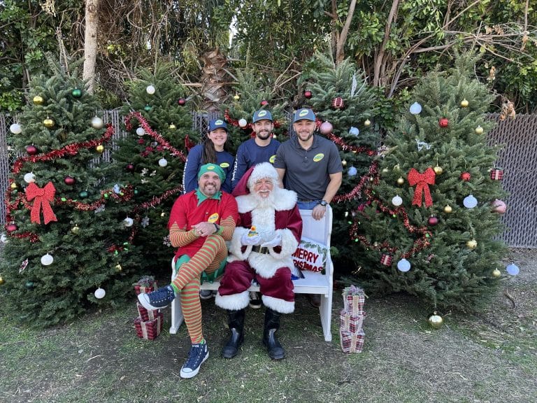 Santa and an elf pose with four staff members wearing caps in front of decorated Christmas trees outdoors.