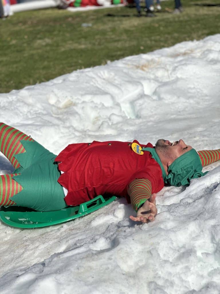 A man in red and green elf costume lies on snow on a green sled, arms outstretched, smiling.