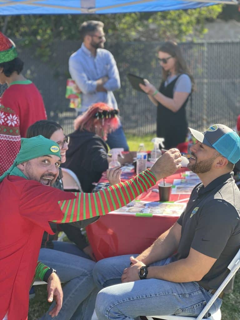A man in green elf hat playfully feeding a smiling man in a blue cap at a festive outdoor holiday table