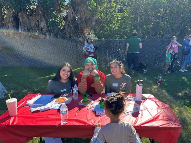 Three smiling volunteers at a red table outdoors helping a child at a community event