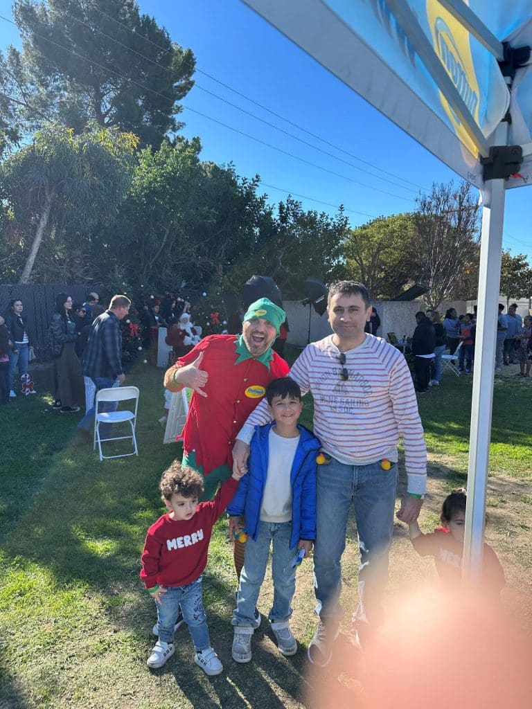 A man and three children pose with a costumed elf at a sunny outdoor holiday event with trees and crowds