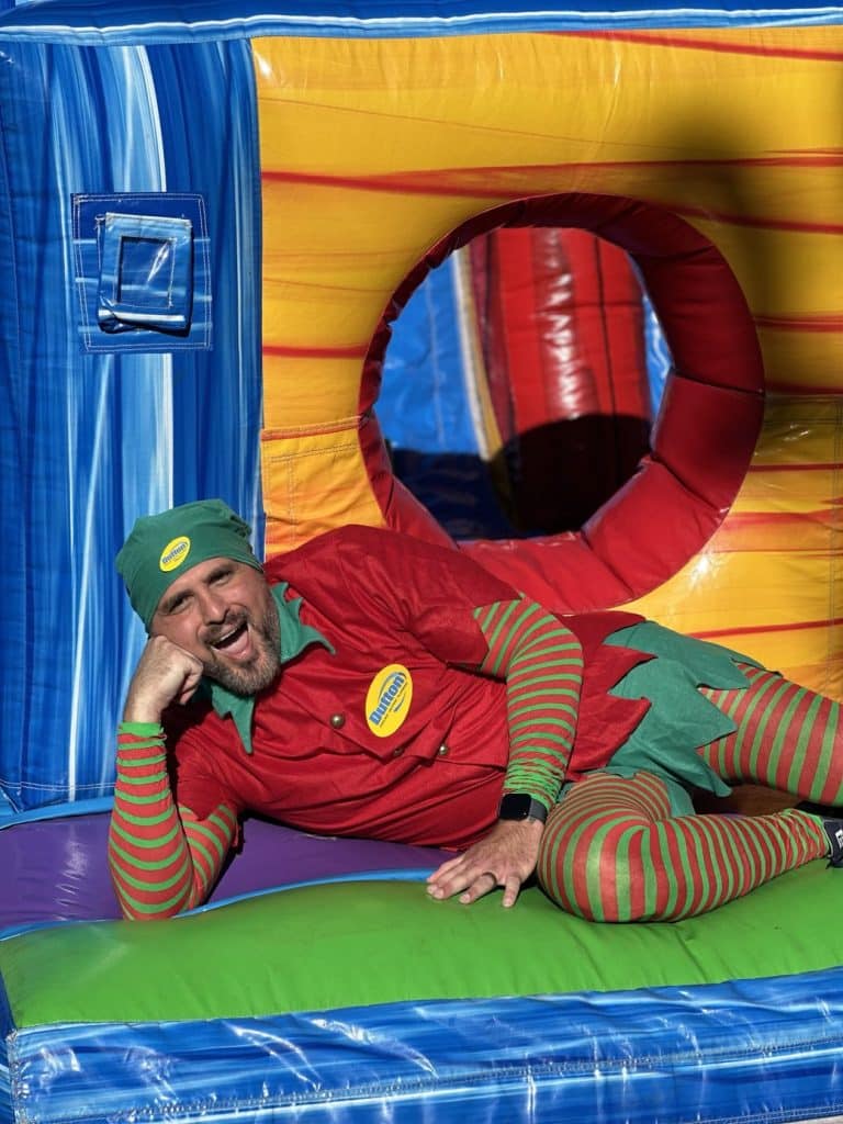 A man in a red and green elf costume lounging and smiling on a colorful inflatable bounce house.