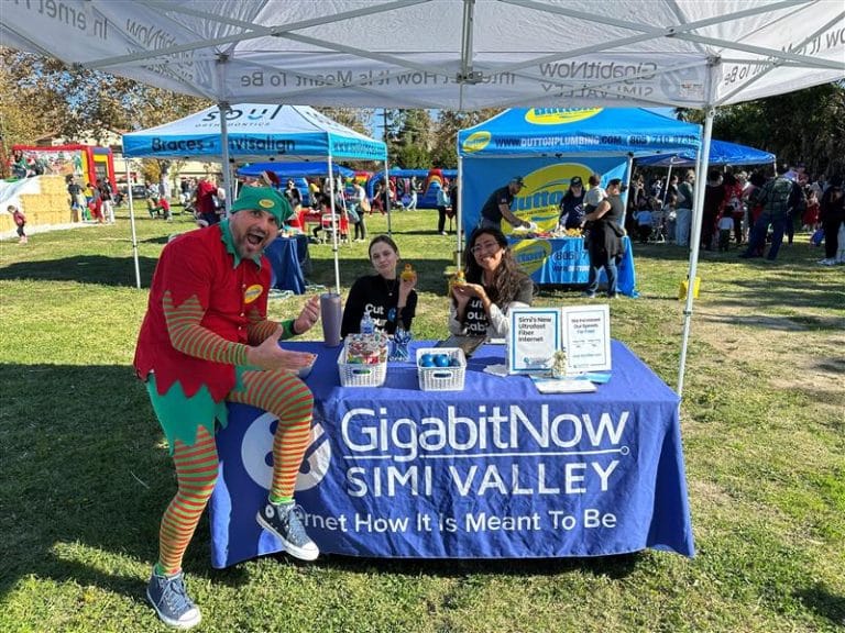 Man in elf costume and two women at GigabitNow Simi Valley booth handing out promotional items at outdoor fair
