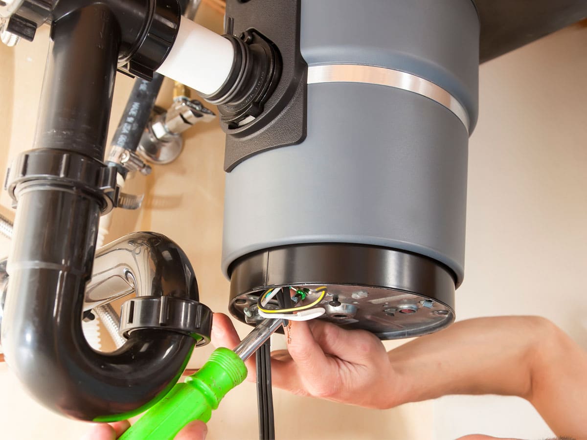 A plumber using a screwdriver to install and wire a garbage disposal under a kitchen sink.