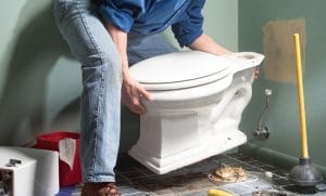 A man lifting and positioning a toilet bowl during bathroom installation with tools and plunger nearby