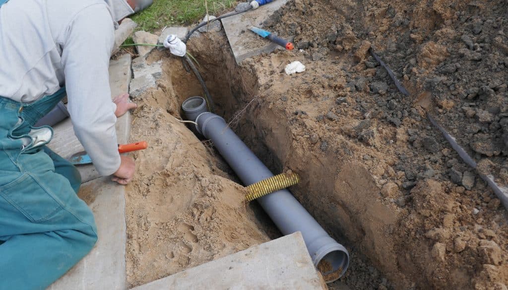 A worker kneeling installs a gray PVC drainage pipe in a sandy trench beside a concrete slab with hand tools.