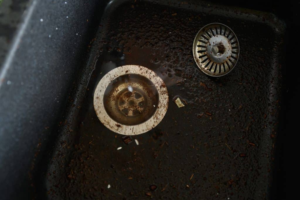 Close-up of a dirty black kitchen sink with coffee grounds, debris, and two metal drain strainers