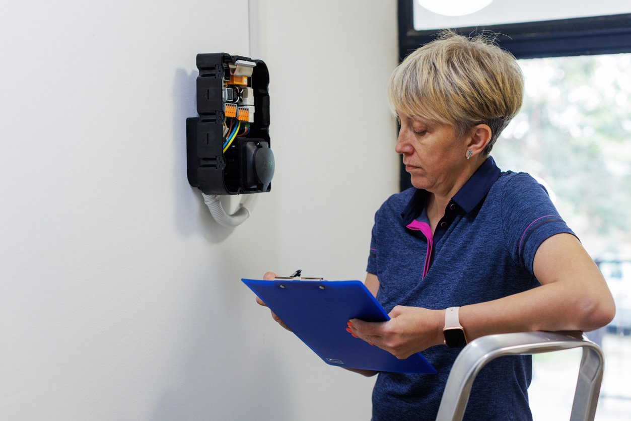 Woman technician inspecting wall-mounted electrical box, taking notes on blue clipboard