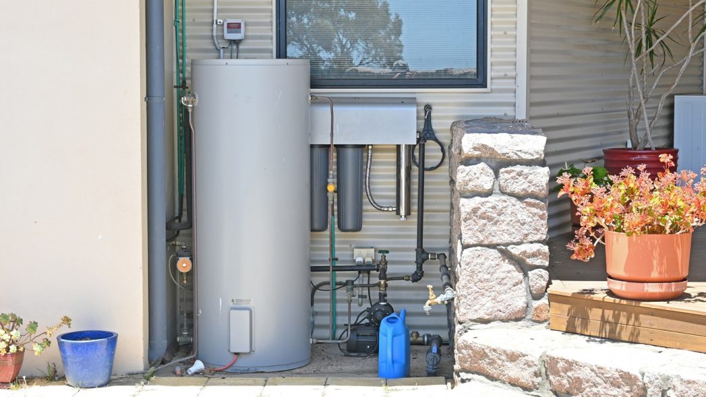 Outdoor water heater and filtration system with pump, pipes, blue watering can and potted plants beside stone pillar