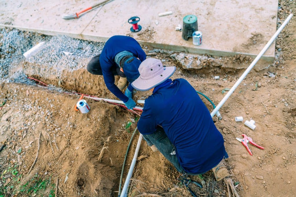 Two construction workers kneeling in a trench installing PVC pipes and wiring near a concrete slab.