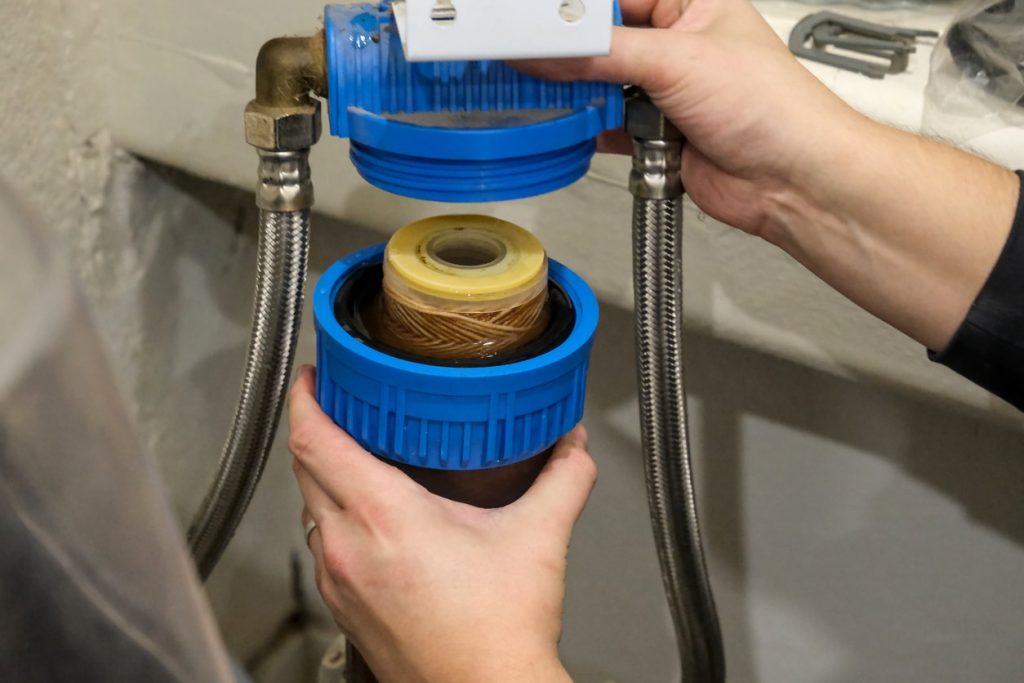 A person replacing an under-sink water filter cartridge in a blue housing with braided stainless supply lines