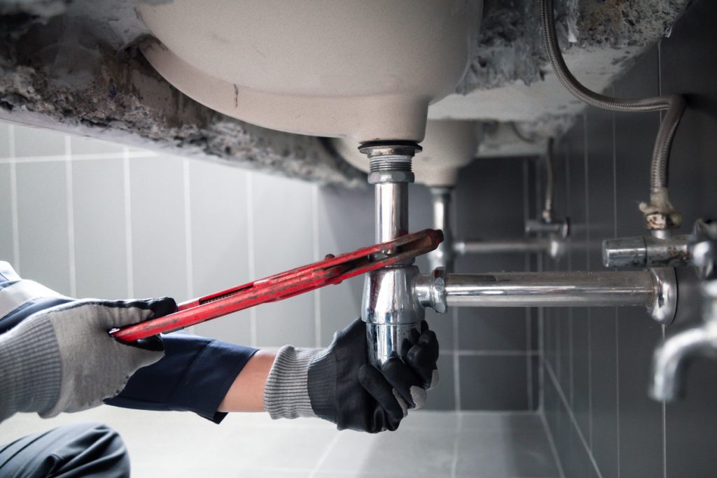 A plumber tightening a chrome P-trap under a sink with a red pipe wrench