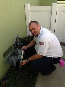 A technician kneeling by an outdoor gas meter using a level, smiling in a white uniform