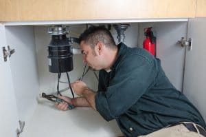 A plumber repairing a garbage disposal under a kitchen sink with pliers.