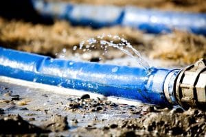 Blue water pipe with a leak at a coupling, spraying water onto muddy ground