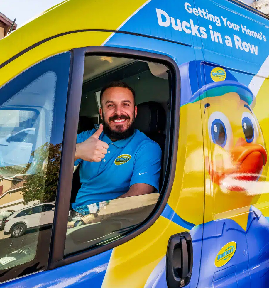A smiling man in a blue polo giving a thumbs-up from a yellow and blue service van with a large duck graphic.